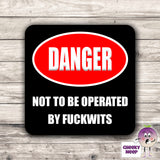Square black hardboard coaster with the words "Danger not to be operated by fuckwits" printed on the coaster. As produced by CheekyNeep.com