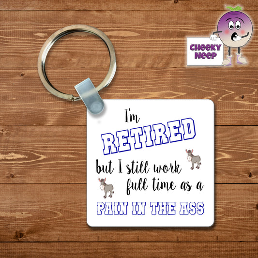 Square plastic keyring with the words "I'm retired but still work full time as a pain in the ass" printed on both sides.